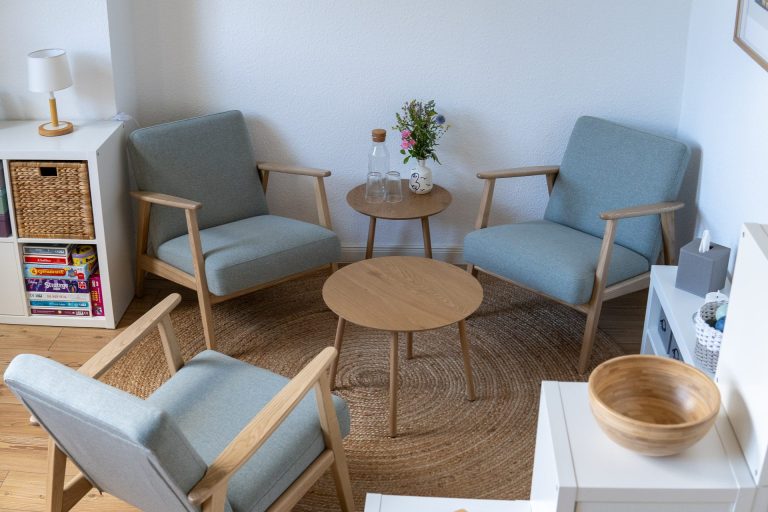 Foto eines Behandlungsraumes: Gemütliche Sessel auf einem weichen Teppich. Große Fenster im Hintergund. Bunte Bilder an der Wand.